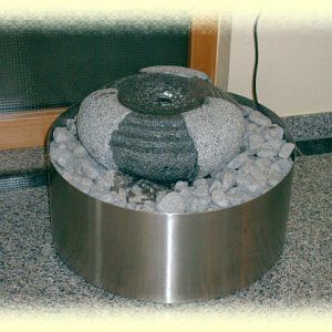 Kaskadenförmig bearbeiteter  Granit-Quellstein auf hellgrauem Zierkies in rundem Edelstahl-Becken       (Durchm.       65, Höhe 25 cm)